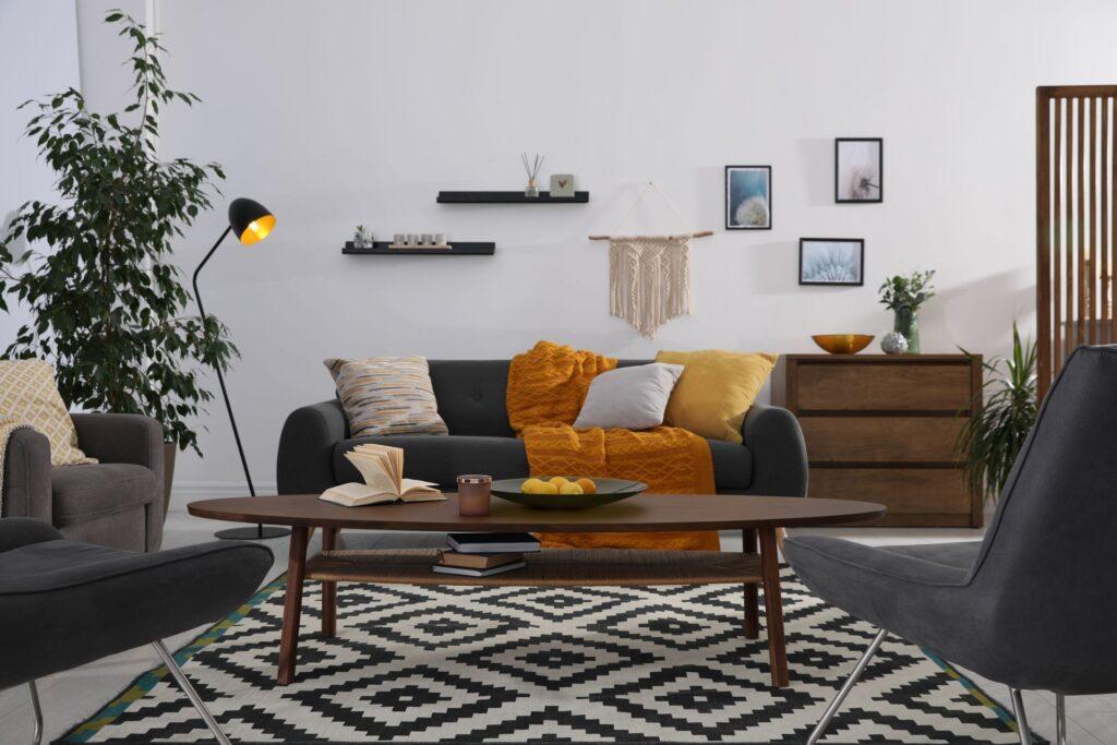 Modernes Wohnzimmer mit grauem Sofa, farbigen Kissen, Holztisch und geometrischem Teppich in schlichter, minimalistischer Einrichtung.